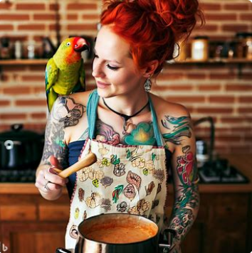 A 29-year-old woman with vibrant red hair tied in a loose, messy bun, wearing a vintage floral apron over a quirky graphic t-shirt that says 'Flavor Philosopher.' She has a twinkle in her green eyes, an infectious smile, and holds a wooden spoon in one hand. Perched on her shoulder is a cheeky parrot named Gumbo, who seems interested in a pot of exotic stew simmering on a stove behind her.